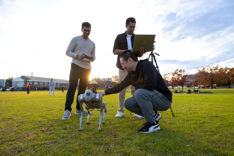 Three students interact with a robotic "dog" in a field with the sun shining through them