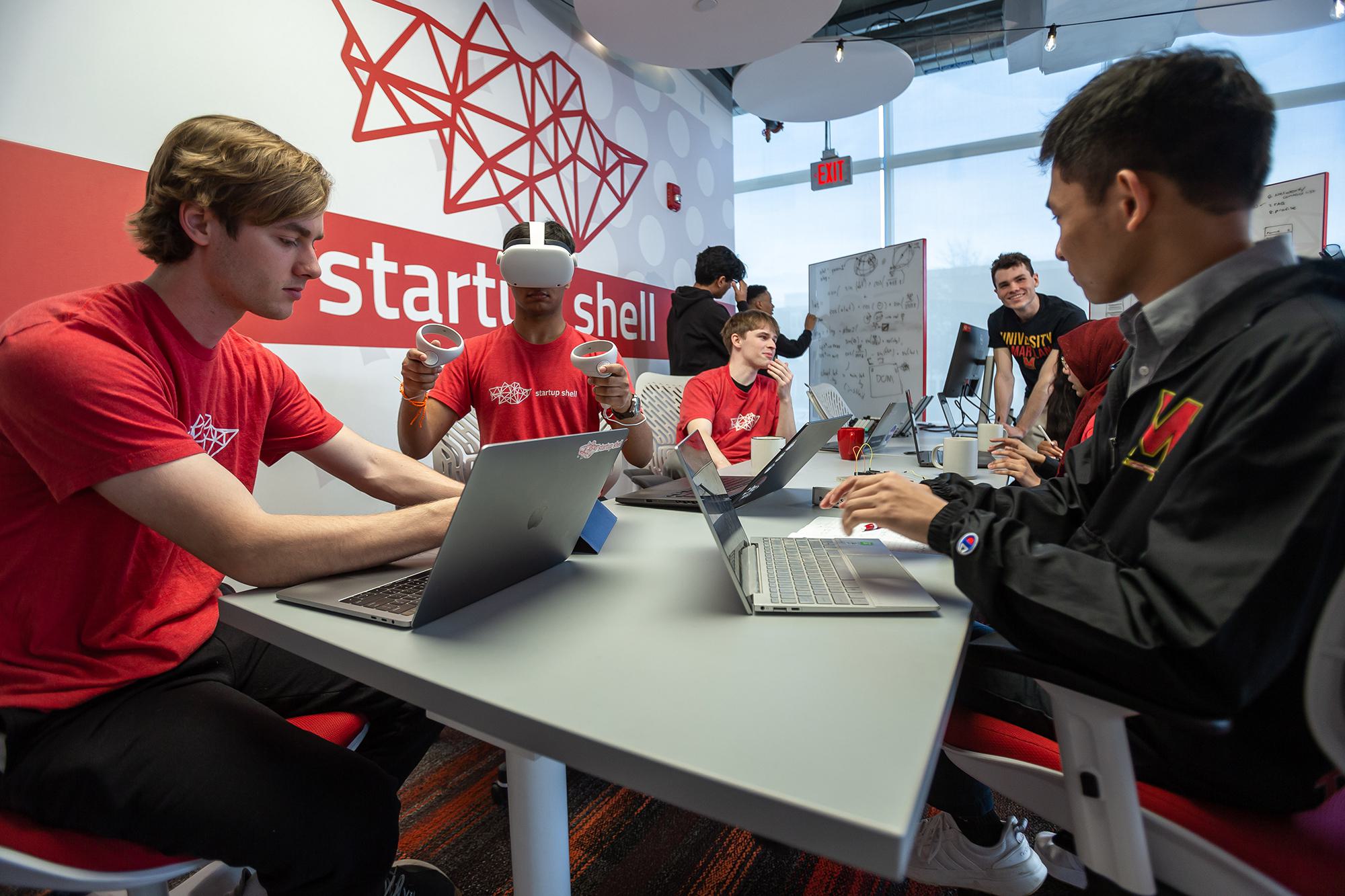 students sitting around a table with laptops. One student has a VR headset on