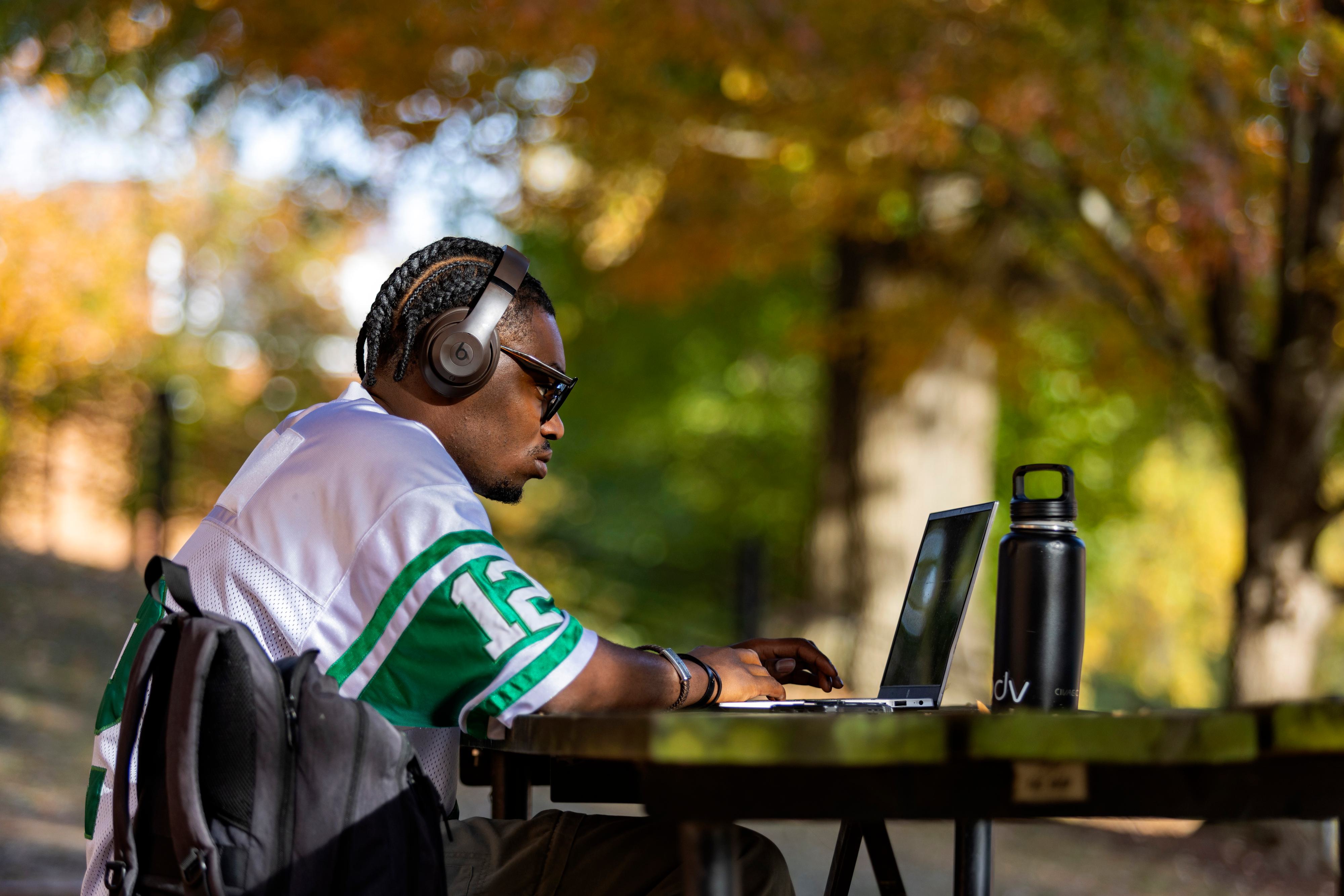 A student sits outside on a fall day working on a laptop