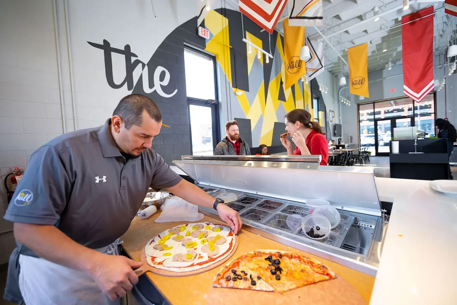 A chef at The Hall CP carefully prepares a pizza on a counter dusted with flour next to a row of ingredients. Restaurant patrons enjoy their meals while having conversations in the background