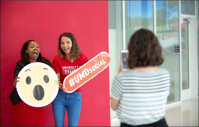 Employees pose with with an emoji surprised face sign and a hashtag UMD social sign as a photographer takes their photo in the foreground