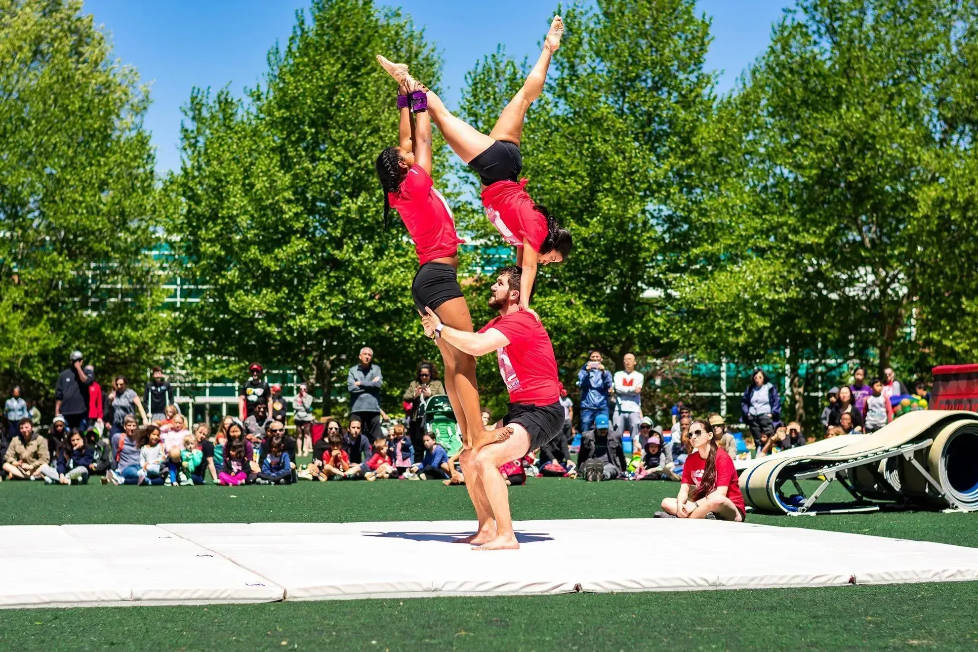 A Gymnastics team of 3 in Maryland Red shirts are stacked in a complicated pose requiring each performer have perfect balance while holding each other. A crowd of spectators watch with anticipation in the background.